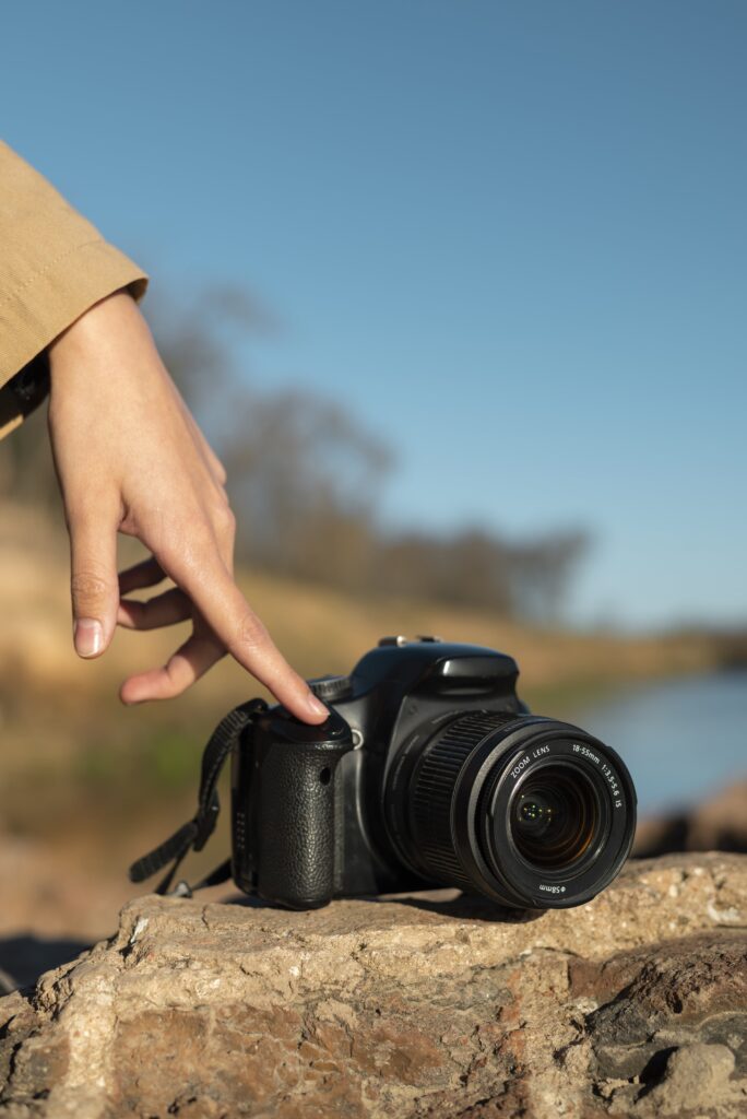 A hand reaching to press the shutter button on a DSLR camera placed on a rock outdoors.
