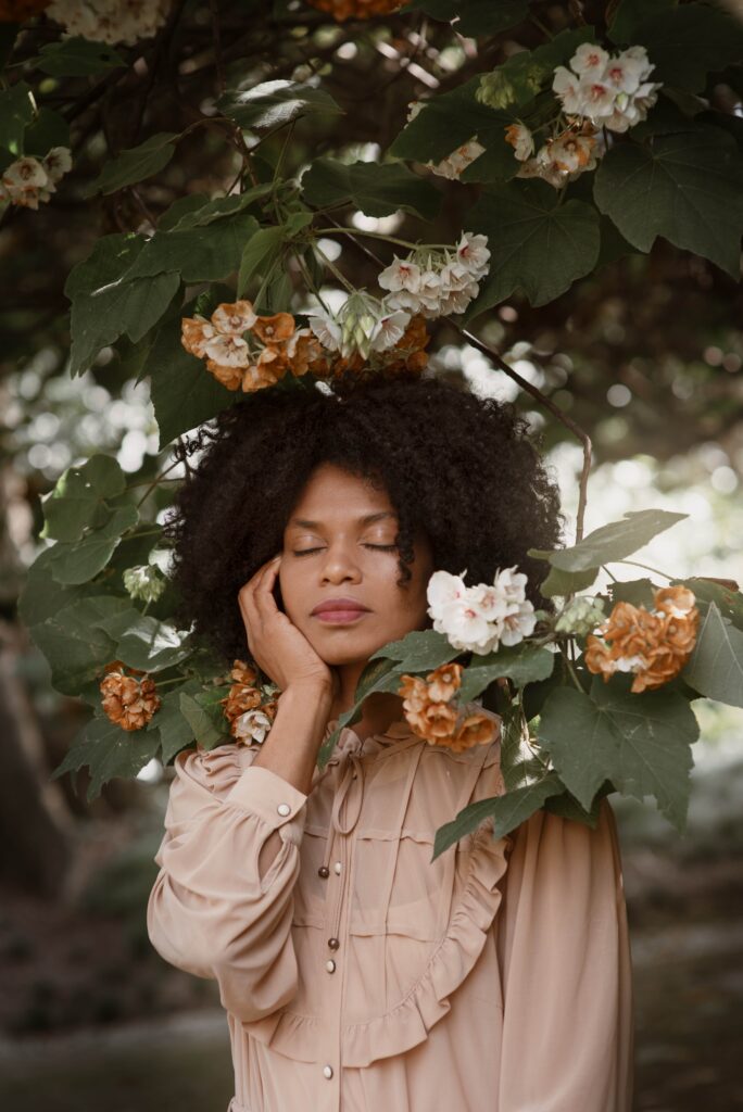Woman posing with her eyes closed, surrounded by flowers and leaves in a garden.