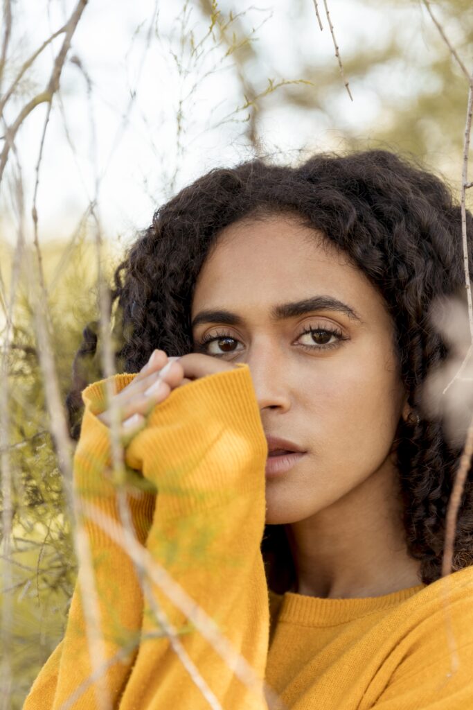 Woman in a yellow sweater looking through branches, with a thoughtful expression.