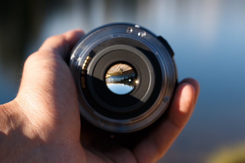 Lens & Light: The Art of Capturing Moments. A close-up of a hand holding a camera lens, with a clear view of a landscape reflected inside the lens.
